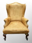 A George III style wingback armchair upholstered in a studded tan faux leather, raised on castors,