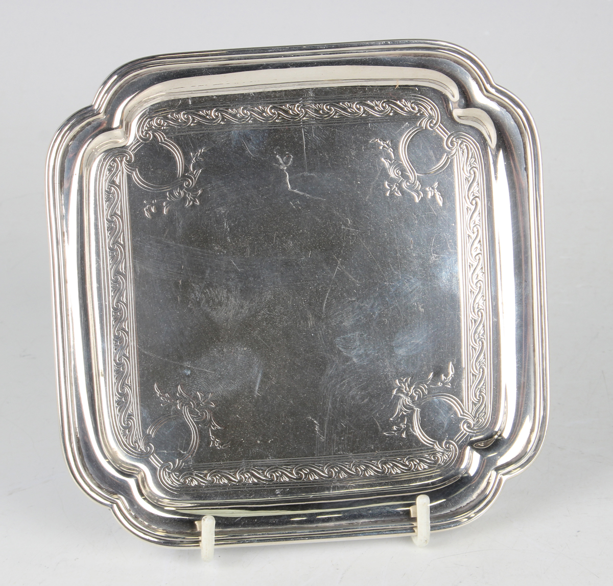 A George V silver card salver of square form with lobed corners, engraved with scrolls, raised on