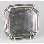 A George V silver card salver of square form with lobed corners, engraved with scrolls, raised on