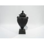 A Wedgwood black basalt lidded urn with ram's head detail. The lid is damaged and nibble to rim,