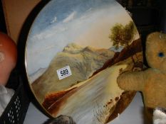 A hand painted wall plaque featuring mountains by Doulton artist M W K Blair, 36 cm diameter.