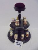 Fifteen Royal Crown Derby thimbles on a thimble stand with pin cushion.
