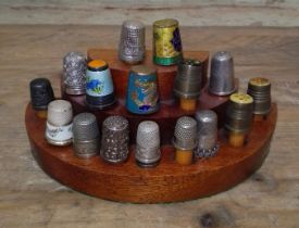 A wooden stand containing various thimbles to include enamelled, silver, white metal, etc.