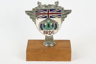 A vintage enamel car grill badge for 'BRDC' mounted to wooden plinth.