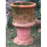 A weathered cylindrical terracotta planter with repeating foliate panels raised on associated