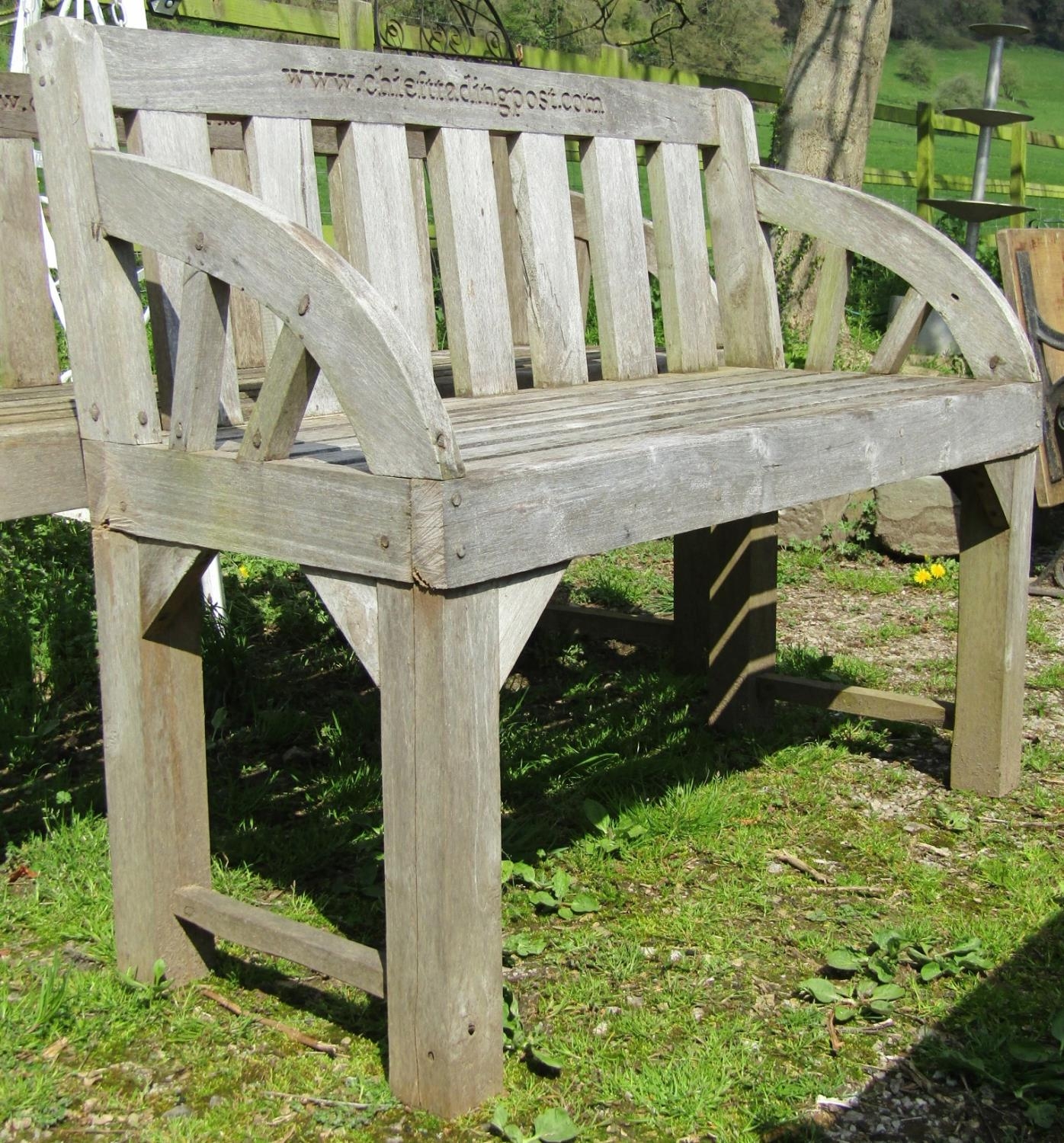 A weathered hardwood two seat garden bench with slatted seat, back and curved arms (stamped to