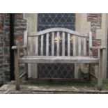 A weathered teak two seat garden bench with slatted seat and back beneath an arched rail 124 cm wide