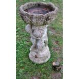 A small weathered cast composition stone bird bath in the form of three cherubs supporting a