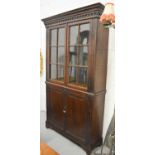 A slim oak cabinet circa 1900, the upper section having a dentil mouled cornice above two glazed