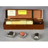 A cylinder slide rule with bakelite handle, boxed and three glass inkwells with bakelite lids.The