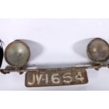 Pair of vintage vehicle headlights, mounted on a bar, with registration plate.