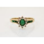 An emerald and diamond yellow metal ring, the central oval cut emerald in a surround of round cut
