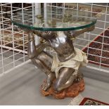 A polychrome wooden side table of a kneeling young man with round clear glass top, after an