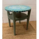 A blue painted circular table with green and blue mosaic top and shelf under