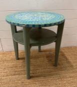 A blue painted circular table with green and blue mosaic top and shelf under
