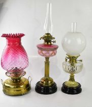 A brass oil lamp with a large cranberry glass shade, together with two further brass oil lamps,