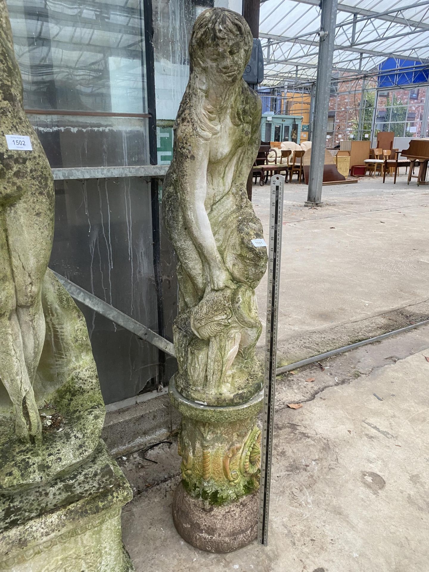 A DECORATIVE RECONSTITUTED STONE FIGURE OF A FEMALE CARRYING A WATER JUG ON A DECORATIVE