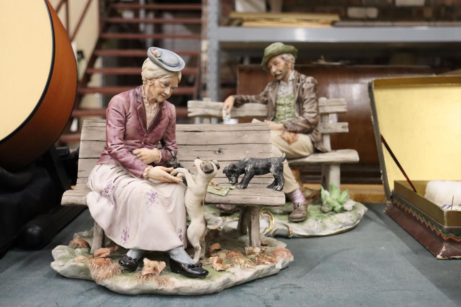 TWO CAPODIMONTE STYLE ORNAMENTS, A MAN ON A BENCH PLAYING CARDS AND A LAWITH DOGSDY ON A BENCH