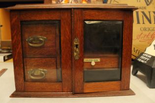 AN OAK TWO GLASS DOOR SMOKERS CABINET WITH THREE INTERIOR DRAWERS ONE LABELED PLAYING CARDS COMPLETE