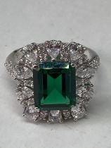 A WHITE METAL RING WITH A LARGE RCTANGULAR LABORATORY EMERALD SURROUNDED BY CLEAR STONES ALSO ON THE