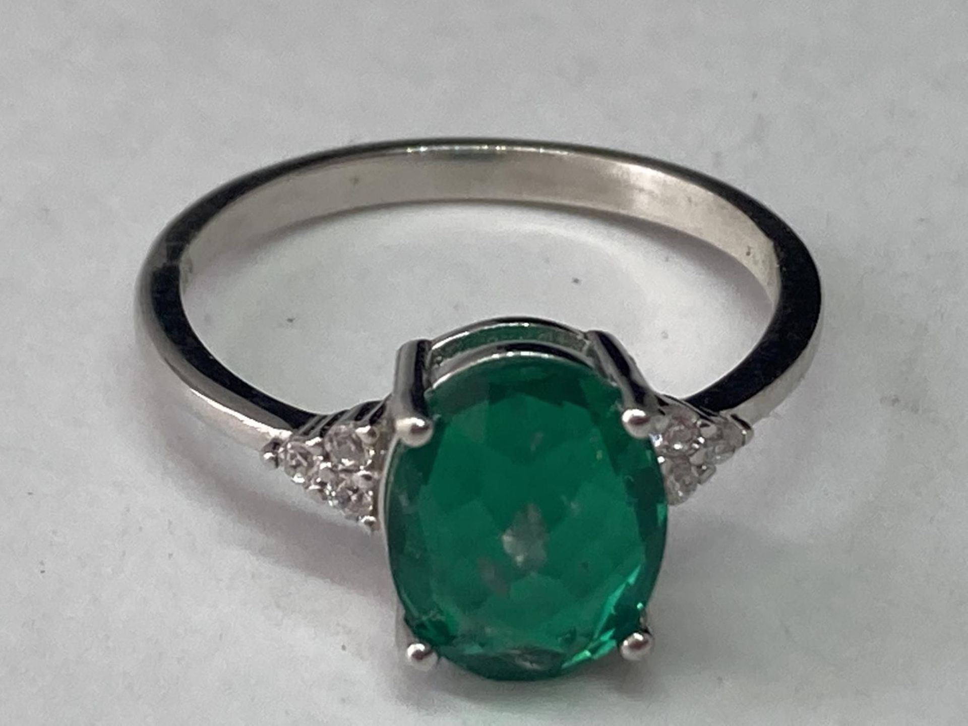 A WHITE METAL RING WITH AN OVAL LABORATORY EMERALD WITH THREE CLEAR STONES TO EACH SIDE SIZE Q/R