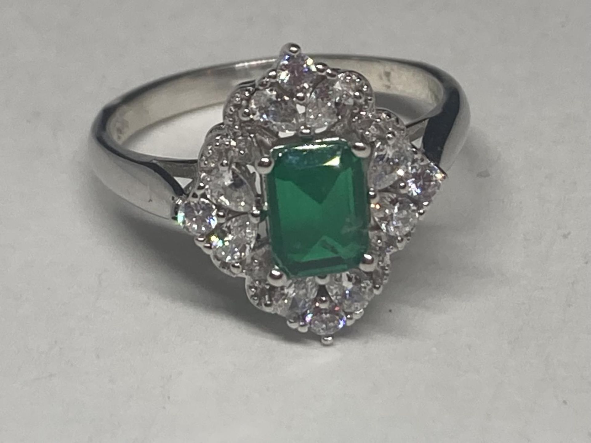 A WHITE METAL RING WITH A RECTANGULAR LABORATORY EMERALD SURROUNDED BY CLEAR STONES SIZE Q/R