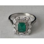 A WHITE METAL RING WITH A CENTRE RECTANGULAR LABORATORY GROWN EMERALD SURROUNDED BY CLEAR STONES