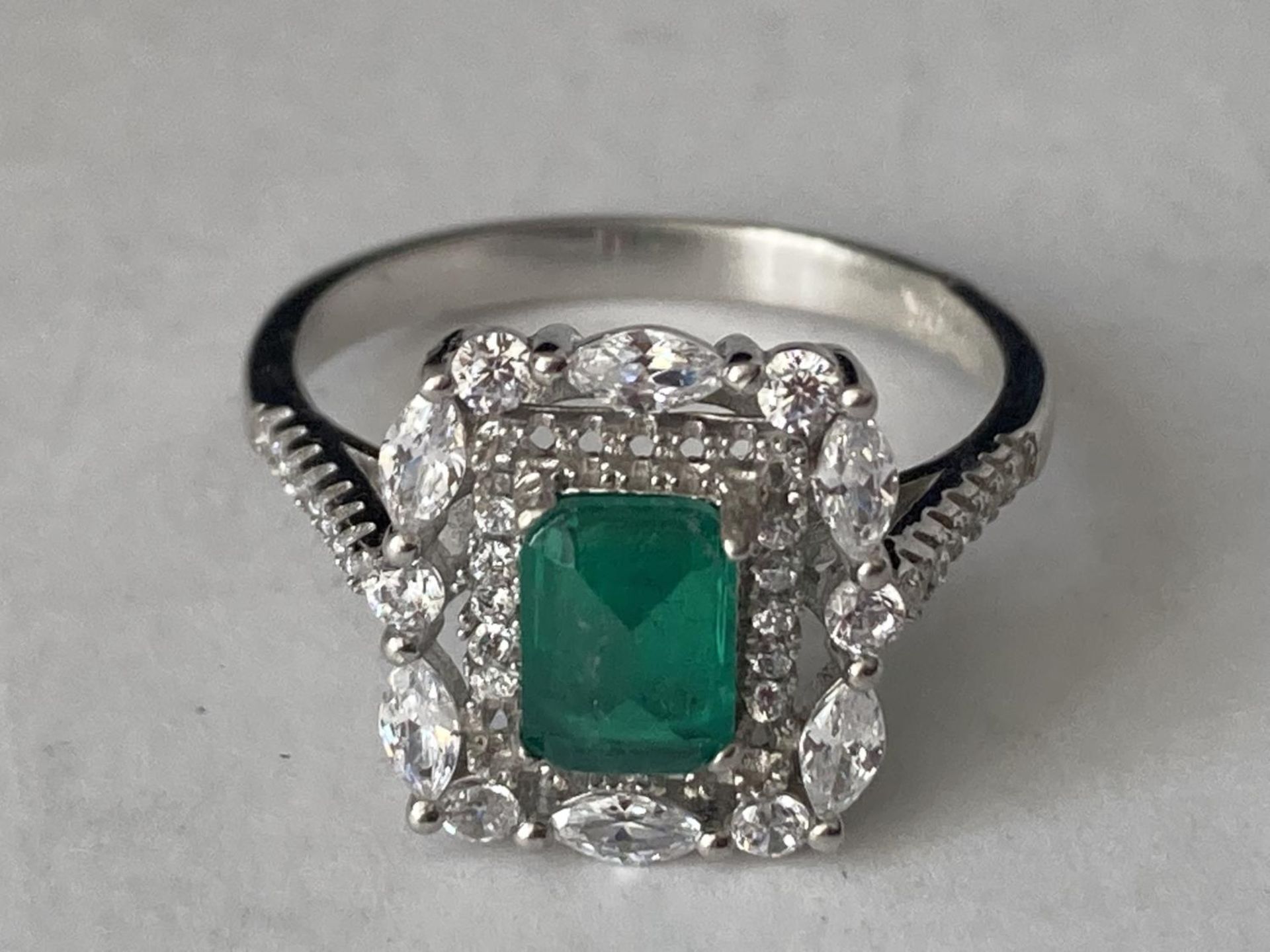 A WHITE METAL RING WITH A CENTRE RECTANGULAR LABORATORY GROWN EMERALD SURROUNDED BY CLEAR STONES