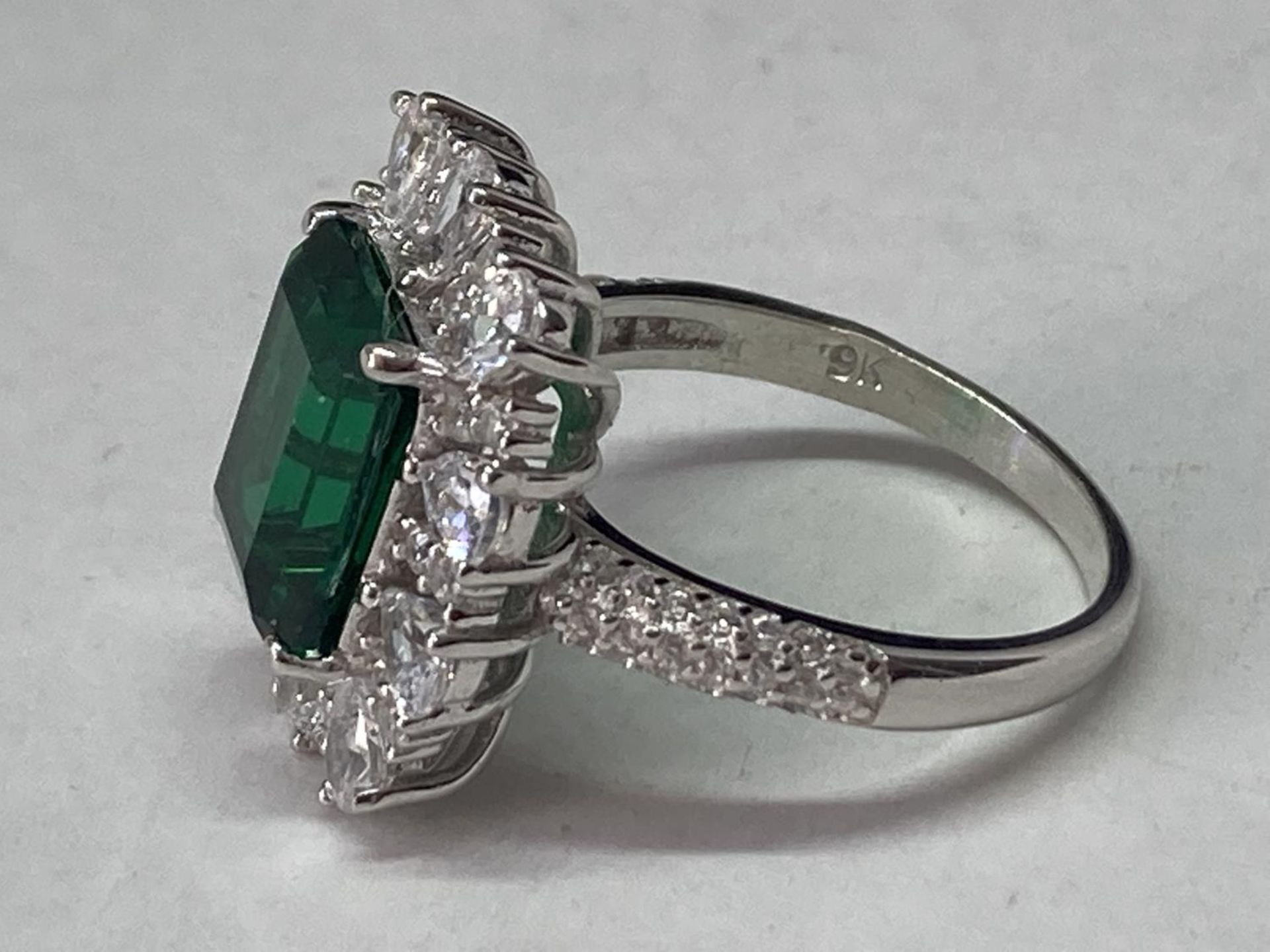 A WHITE METAL RING WITH A LARGE RCTANGULAR LABORATORY EMERALD SURROUNDED BY CLEAR STONES ALSO ON THE - Image 3 of 8