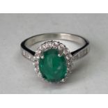 A WHITE METAL RING WITH A CENTRE LABORATORY GROWN EMERALD WITH CLEAR STONES SURROUNDING AND ON THE