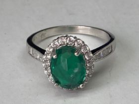 A WHITE METAL RING WITH A CENTRE LABORATORY GROWN EMERALD WITH CLEAR STONES SURROUNDING AND ON THE