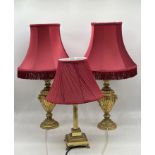 A pair of gilt lamps along with a brass column lamp, all with maroon shades.