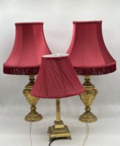 A pair of gilt lamps along with a brass column lamp, all with maroon shades.