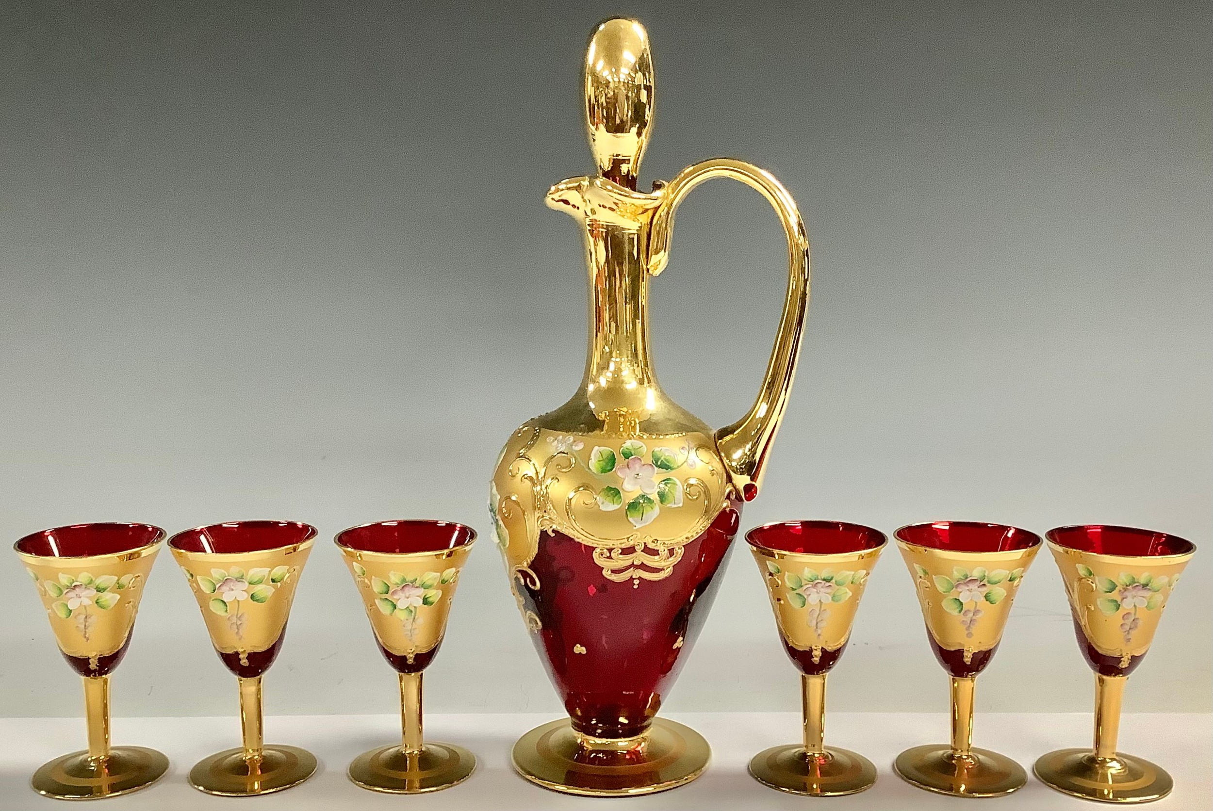 A Murano ruby glass decanter and six wine glasses, gilded and painted with flowers in relief; a