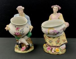 A pair of Sevres style figural spill holders, as Dandy and companion each holding floral bowls, faux