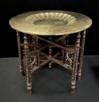 A Middle eastern brass top table with carved and pierced wooden supports, the brass charger top with