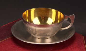 A Russian silver tea cup and saucer, engraved with a topographical view, on a textured ground,