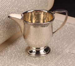 A E Jones - an Arts and Crafts silver pedestal cream jug, rope-twist border, planished overall,