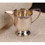 A E Jones - an Arts and Crafts silver pedestal cream jug, rope-twist border, planished overall,