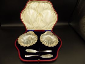 A pair of cased silver scallop shell shaped butter dishes and knives. Hallmarked - Sheffield 1901 by
