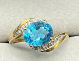 9ct yellow gold ring set with an oval cut blue topaz stone off set with diamond shoulders. [Ring