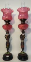 A Pair of Egyptian Revival figural oil lamps set with Victorian cranberry glass shades [72cm]