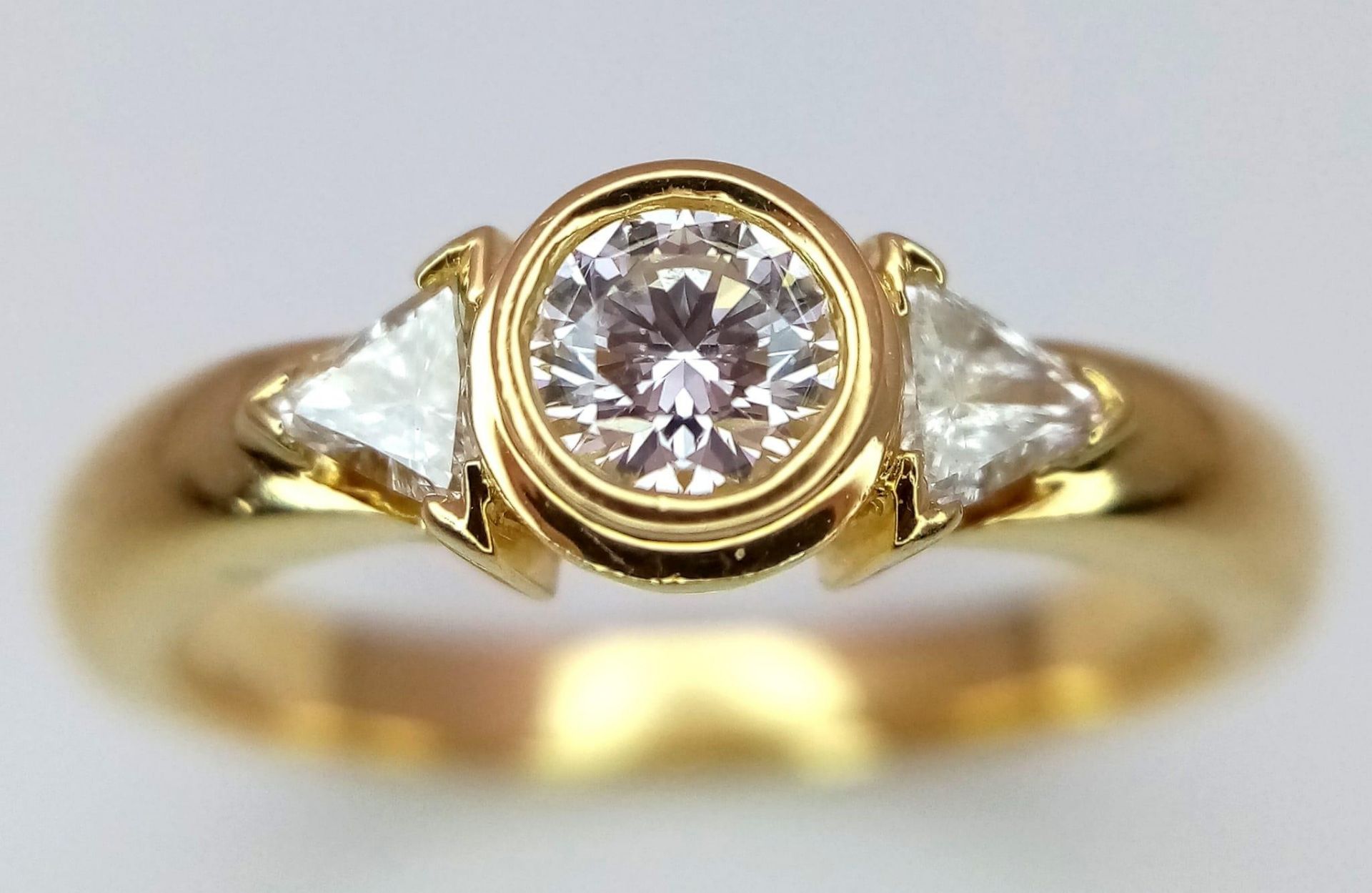 An 18K Yellow Gold Diamond Ring. Central round cut diamond with trillion cut diamond accents. Size