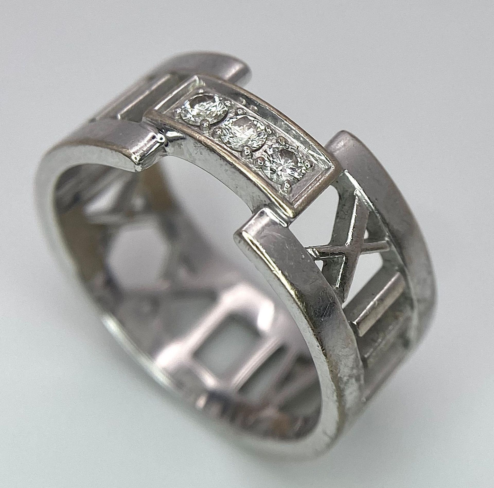 An 18K White Gold Tiffany Atlas Diamond Ring. Pierced Roman numeral decoration. Tiffany mark. Size
