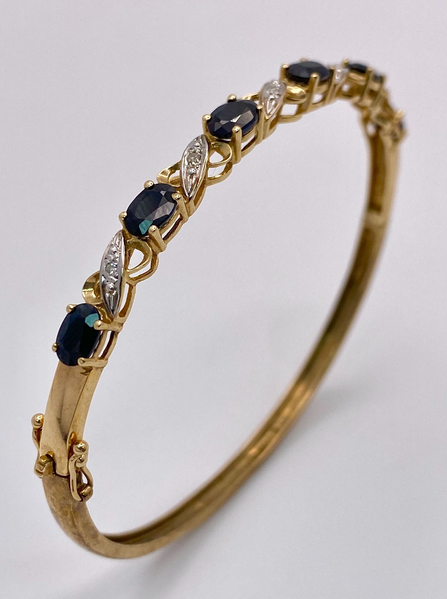 A Vintage 9K Yellow Gold Sapphire and Diamond Bangle. Six oval cut sapphires with diagonal