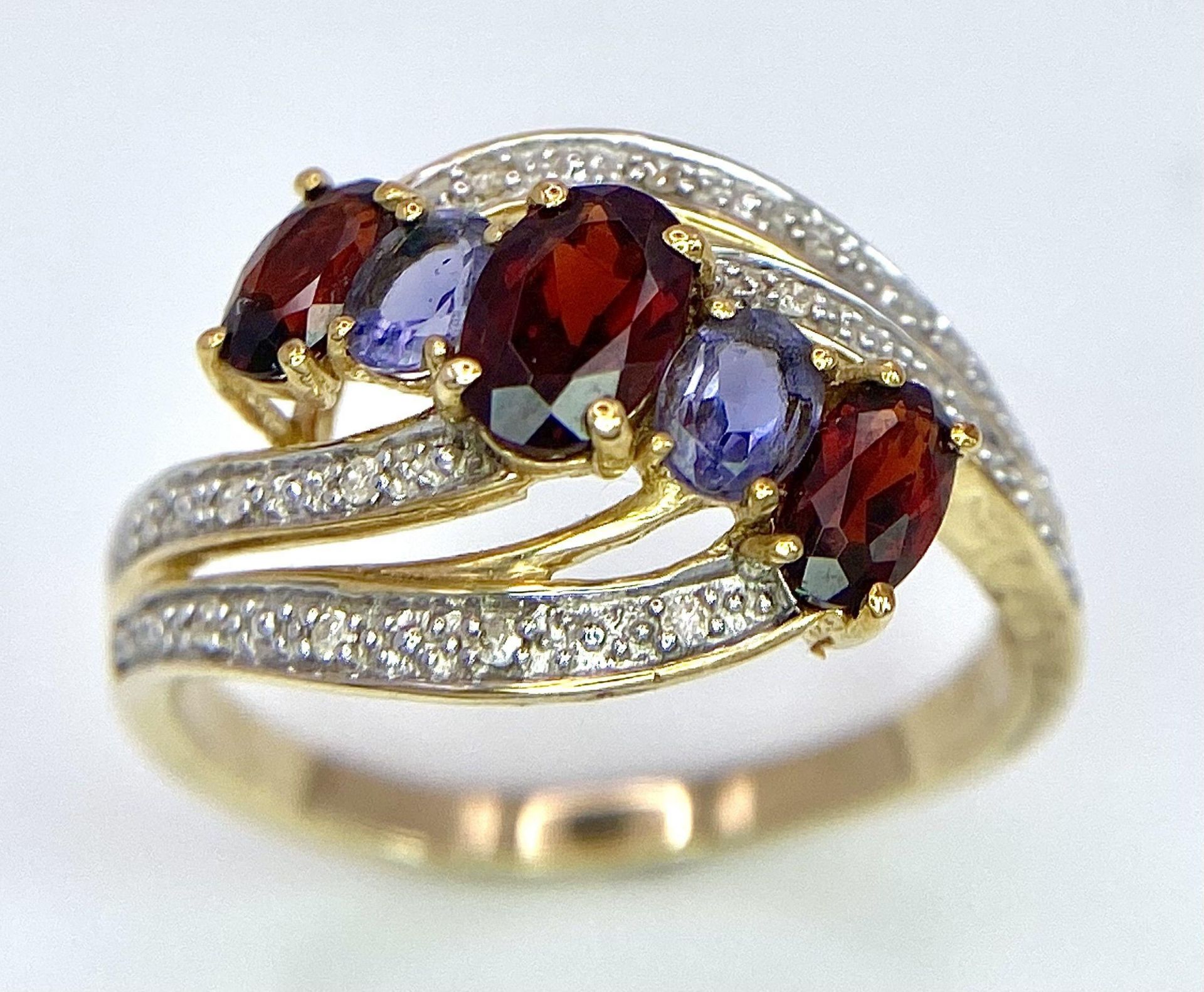 A 9 Carat Yellow Gold Garnet, Tanzanite and Diamond Set Crossover Ring Size P. Crown Measures 1.