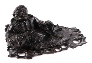 A Chinese hardwood figure, of a recumbent bearded man holding a bat seated beside a temple lion,