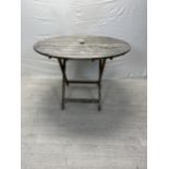 A vintage weathered teak folding garden table. H.75cm Dia.110cm.