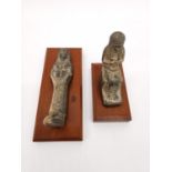 Two Egyptian-style clay ushabti on wooden plinth bases. One lying down and one seated on a