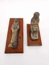 Two Egyptian-style clay ushabti on wooden plinth bases. One lying down and one seated on a